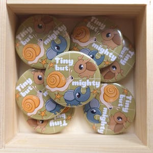 May include: A collection of round pinback buttons featuring a light olive green background with a white grid pattern. Each button displays a cartoon snail, a beetle, and a stag beetle with the text Tiny but mighty and small orange star accents.