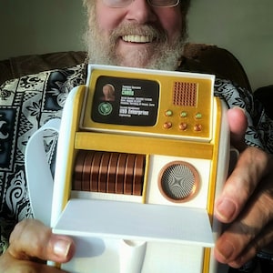 May include: A gold and white Star Trek prop replica, featuring a screen with text and a series of copper-colored discs. The prop has a retro design with a grid-like speaker and several round buttons. The text on the screen reads "USS Enterprise".