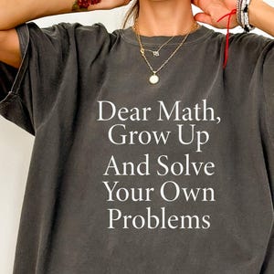 Könnte beinhalten: Dunkelgraues T-Shirt mit dem weißen Text "Dear Math, Grow Up And Solve Your Own Problems". Das T-Shirt hat einen Rundhalsausschnitt und eine lockere Passform. Das Wort "PEPPER" befindet sich in einem Kreis unten links.