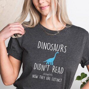 May include: A dark gray t-shirt with a blue dinosaur illustration and the text "Dinosaurs Didn't Read Now They Are Extinct".