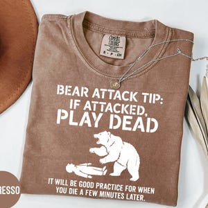 May include: An espresso-colored short-sleeve t-shirt featuring white text that reads: BEAR ATTACK TIP: IF ATTACKED, PLAY DEAD. IT WILL BE GOOD PRACTICE FOR WHEN YOU DIE A FEW MINUTES LATER. A white graphic of a bear standing over a person is included.