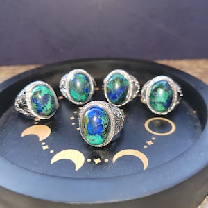 Op de afbeelding: Vijf zilveren ringen met ovale, groen-blauwe edelsteen bezettingen. De ringen worden gepresenteerd op een zwarte dienblad met een halve maan ontwerp.