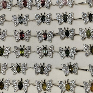 May include: A collection of silver butterfly rings with various colored gemstones. The rings are arranged in rows on a white surface.
