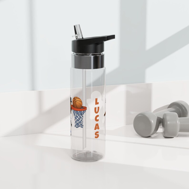 May include: A clear plastic water bottle with a black lid and a straw. The bottle has a basketball and hoop design with the name "Lucas" printed on the side.