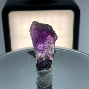 May include: A purple amethyst crystal with a rough, natural shape. The crystal is sitting on a white surface with a reflection of the crystal in the surface.