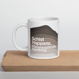 May include: White ceramic mug with a brown and gray abstract design. The mug has the text "Schist Happens. Metamorphically Speaking." printed on it.