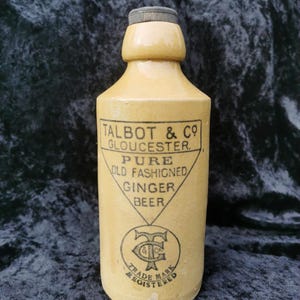 May include: A tan stoneware ginger beer bottle with a dark grey cap. The front features black text reading TALBOT & CO GLOUCESTER PURE OLD FASHIONED GINGER BEER inside a triangle, with a circular logo below containing the letters T & C and the words TRADE MARK REGISTERED.