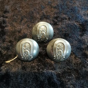 May include: Three circular silver-toned metal buttons, each featuring a raised embossed design of a horseshoe surrounding a central starburst motif. The buttons have a smooth, polished finish and a slightly domed shape, with a small white thread attached.