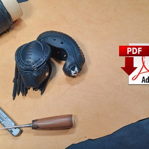 Op de afbeelding: Een zwart leren Predator-masker met ingewikkelde stiksels en franjes, samen met leerbewerkingsgereedschap en een spoel zwart garen. Een PDF-pictogram met het Adobe-logo is ook aanwezig, wat wijst op een digitaal patroon.