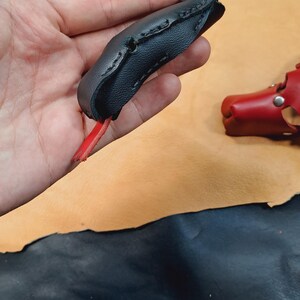 May include: A black leather finger guard featuring a curved, stitched design with a loop at the top and a small red leather tab extending from the bottom. The item is constructed from textured black leather with visible dark stitching along the edges.