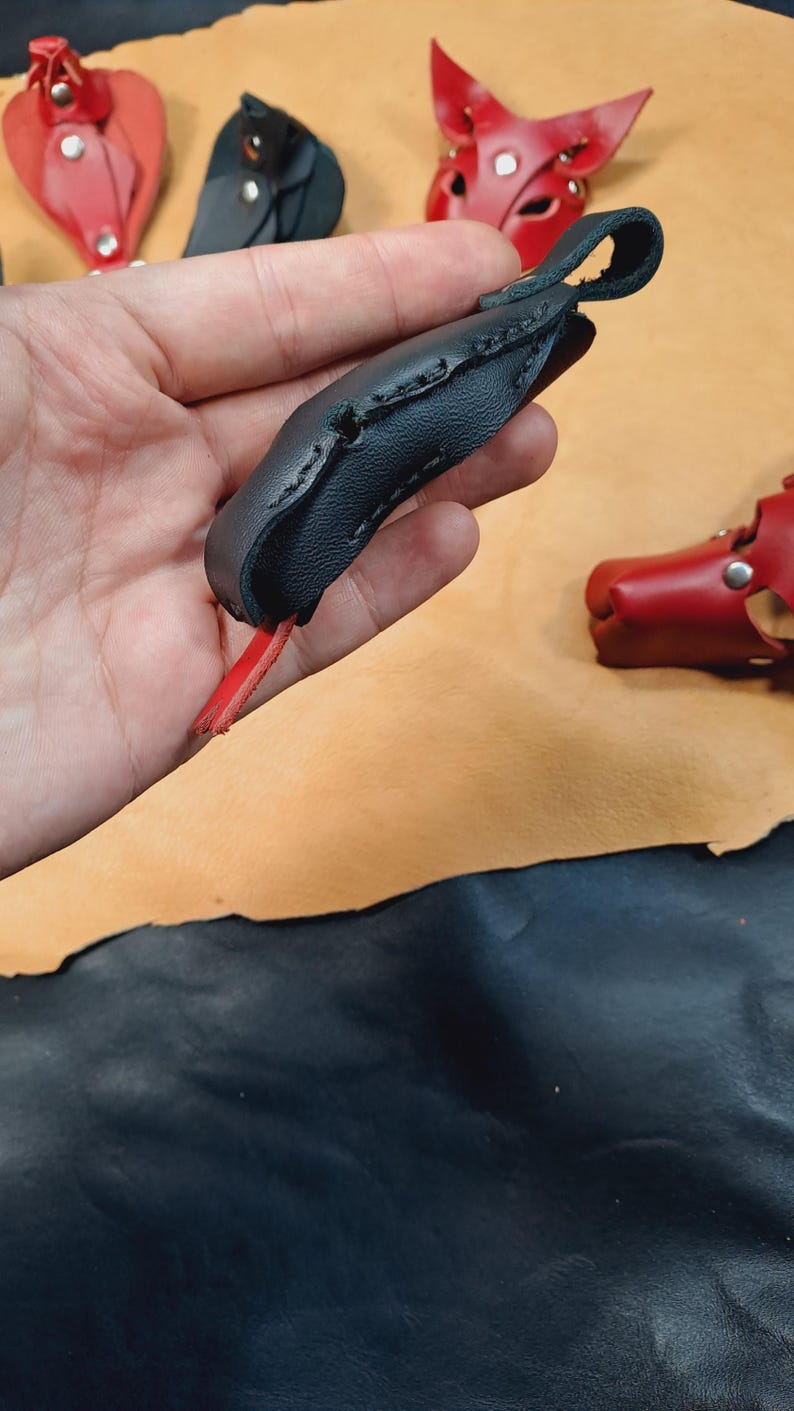 May include: A black leather finger guard featuring a curved, stitched design with a loop at the top and a small red leather tab extending from the bottom. The item is constructed from textured black leather with visible dark stitching along the edges.