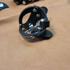 May include: Two black leather circular accessories featuring silver-toned metal rivets and a central silver-toned metal swivel clasp. The leather pieces are constructed with cut-out sections and secured with metal hardware, showcasing a structured design.