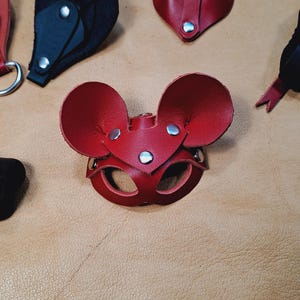 May include: A red leather mouse mask with large rounded ears and silver-toned metal rivets. The mask features two eye openings and is constructed from layered leather pieces. Additional leather mask components in red and black are visible.