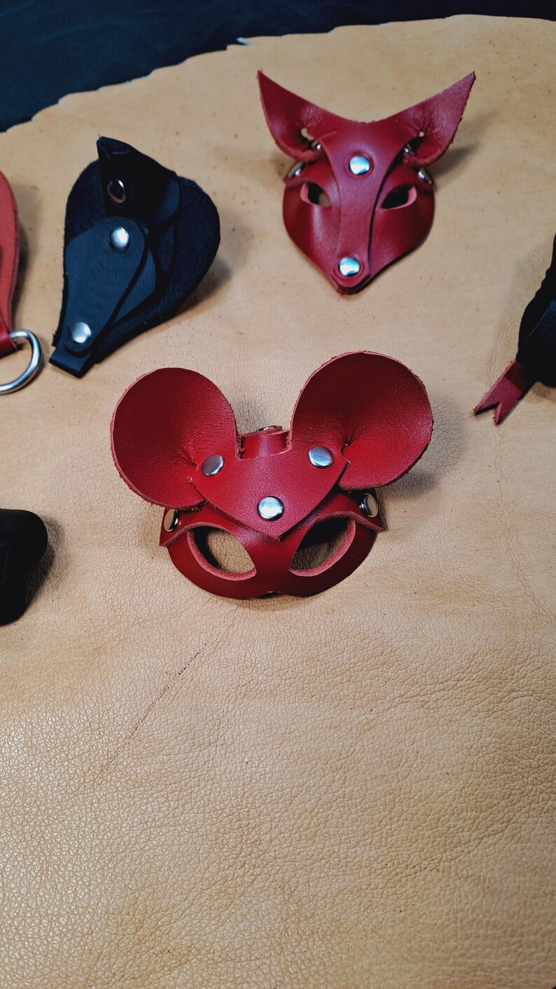 May include: A red leather mouse mask with large rounded ears and silver-toned metal rivets. The mask features two eye openings and is constructed from layered leather pieces. Additional leather mask components in red and black are visible.