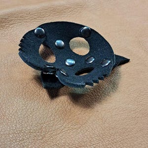 May include: A black leather accessory shaped like a skull with cut-out eye and nose sockets, secured with silver-toned metal rivets. The piece features jagged edges and is displayed alongside other black leather components with metal hardware.