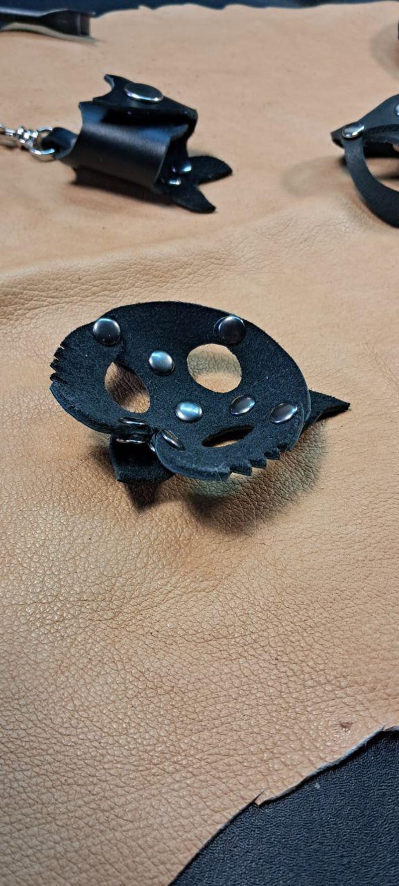 May include: A black leather accessory shaped like a skull with cut-out eye and nose sockets, secured with silver-toned metal rivets. The piece features jagged edges and is displayed alongside other black leather components with metal hardware.
