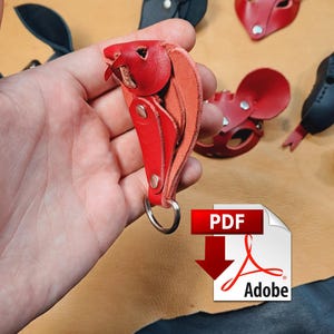 May include: A red leather keyring shaped like a small animal head with a silver metal ring. The keyring is constructed from layered red leather pieces secured with silver metal rivets. Other red and black leather animal masks are visible.