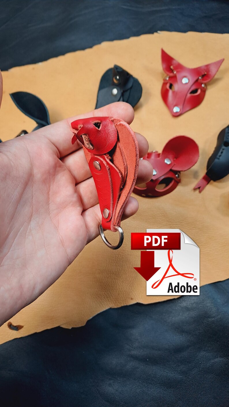 May include: A red leather keyring shaped like a small animal head with a silver metal ring. The keyring is constructed from layered red leather pieces secured with silver metal rivets. Other red and black leather animal masks are visible.