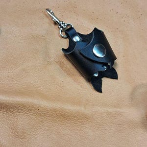 May include: A black leather keychain holder with a silver metal snap button and a swivel lobster clasp. The holder features a cylindrical design with a pointed bottom edge, constructed from smooth black leather with visible stitching.