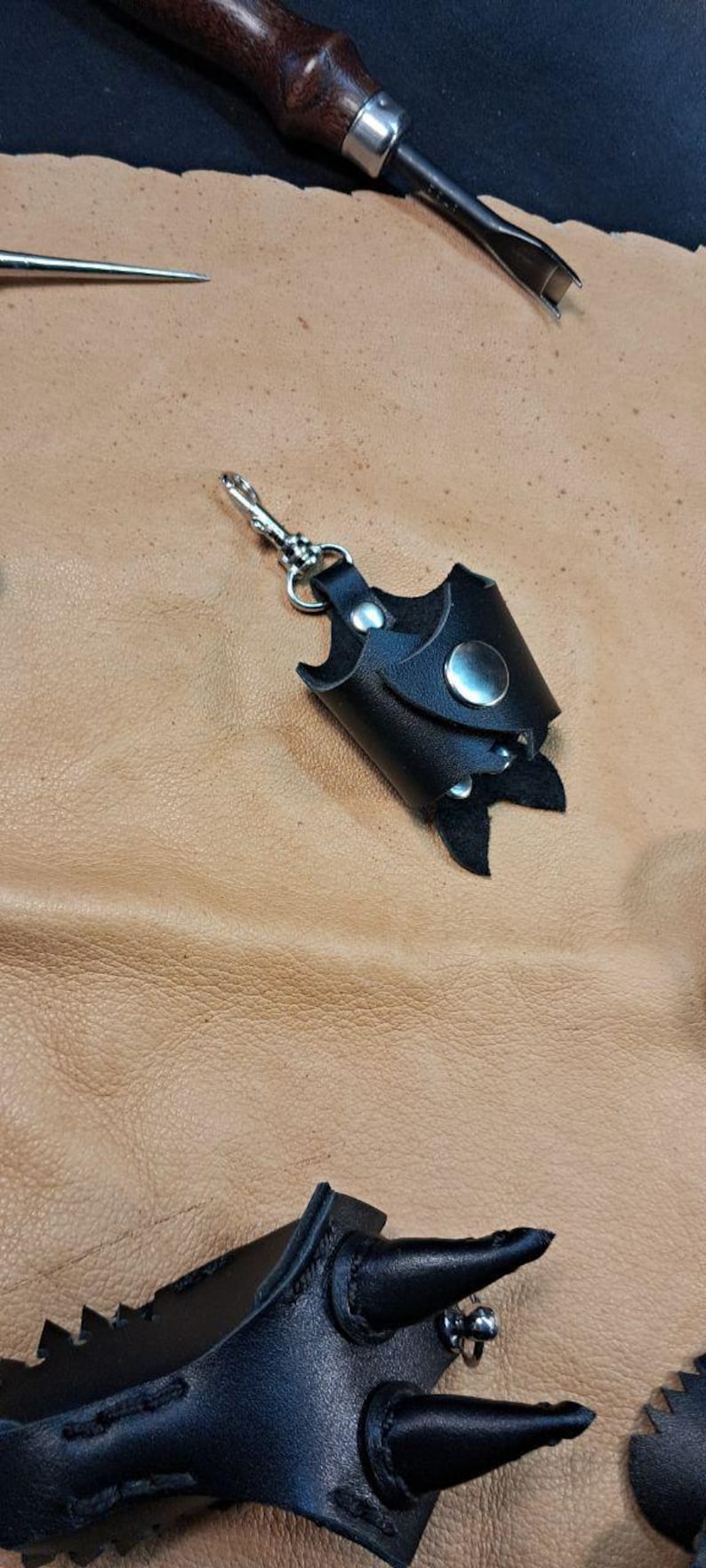 May include: A black leather keychain holder with a silver metal snap button and a swivel lobster clasp. The holder features a cylindrical design with a pointed bottom edge, constructed from smooth black leather with visible stitching.