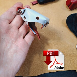 May include: A white leather snake-shaped keyring with silver-tone metal rivets and a red leather tongue. The keyring features a split ring attachment and is displayed alongside other leather animal-shaped keyrings in red and black.