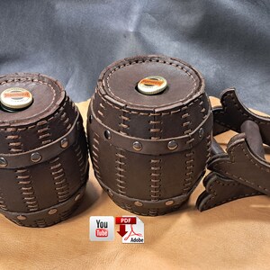 May include: Two brown leather barrels with stitched details and metal accents, one is smaller than the other and both are sitting on a brown surface. A third brown leather object with stitched details and metal accents is to the right of the barrels.