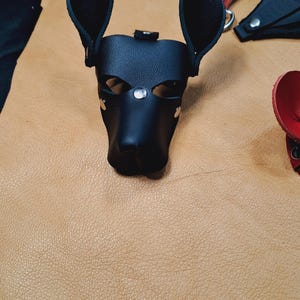 May include: A black leather dog-shaped mask featuring pointed ears, eye cutouts, and silver-toned metal rivets. The mask is constructed from smooth black material with visible stitching and a structured snout area, designed to resemble a canine head.