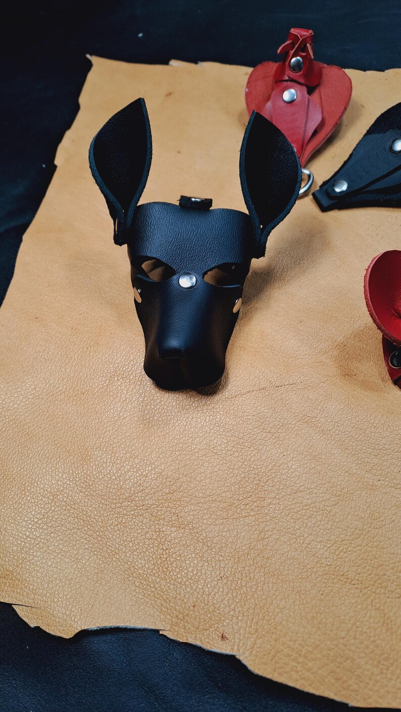 May include: A black leather dog-shaped mask featuring pointed ears, eye cutouts, and silver-toned metal rivets. The mask is constructed from smooth black material with visible stitching and a structured snout area, designed to resemble a canine head.