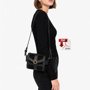 May include: A black shoulder bag featuring a structured cage-like overlay design with a central circular brass-toned ring closure. The bag includes a thin adjustable black shoulder strap and is worn against a black long-sleeved ribbed top.