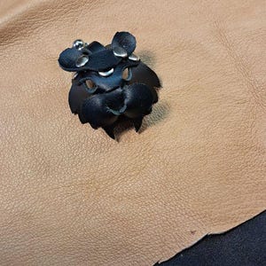 May include: A black leather accessory featuring a rounded shape with cutouts, secured by silver-toned metal rivets. The item has a small, protruding central section and a metallic snap fastener, constructed from layered black leather pieces.