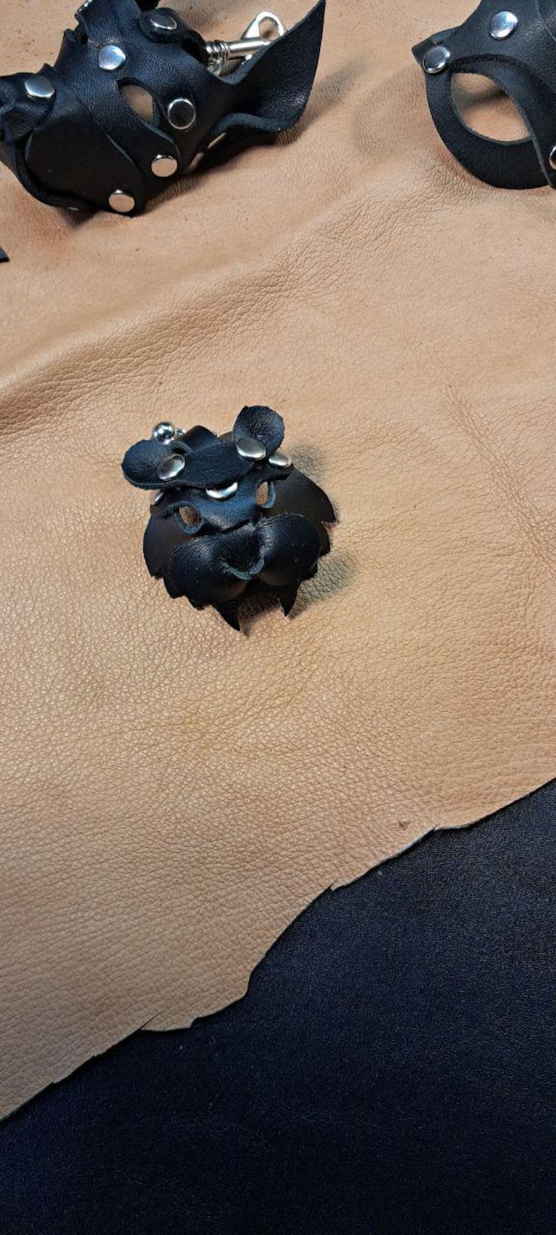 May include: A black leather accessory featuring a rounded shape with cutouts, secured by silver-toned metal rivets. The item has a small, protruding central section and a metallic snap fastener, constructed from layered black leather pieces.