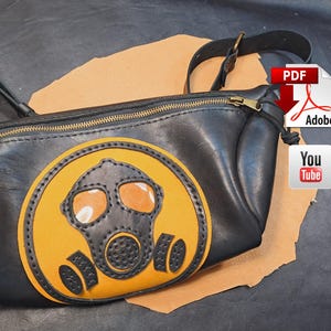 Puede incluir: Riñonera de cuero negro con un diseño de máscara de gas amarilla y negra. La bolsa tiene una cremallera y una correa doradas. El diseño de la máscara de gas está centrado en la parte delantera.