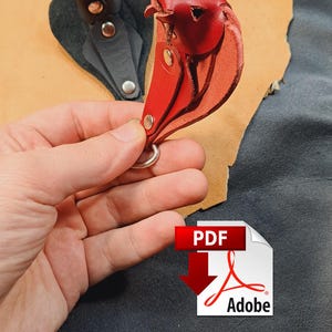 May include: A red leather keyring shaped like a bird, featuring silver-toned metal rivets and a circular metal ring. The keyring is constructed from layered leather pieces, showcasing a smooth finish and visible stitching details.