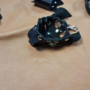 May include: Two black leather finger guards featuring silver-toned metal rivets and snap closures. Each guard has a small metal swivel clasp attached to the side, designed for securing the leather piece around a finger with adjustable metal hardware.
