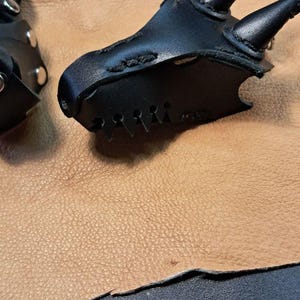 May include: A black leather accessory shaped like a dragon head with two prominent horns, stitched edges, and a metallic snap closure. The item features a pointed snout and a textured surface, presented alongside a matching leather component.