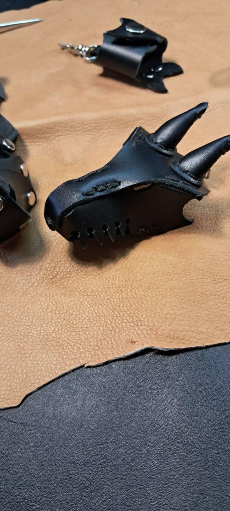 May include: A black leather accessory shaped like a dragon head with two prominent horns, stitched edges, and a metallic snap closure. The item features a pointed snout and a textured surface, presented alongside a matching leather component.