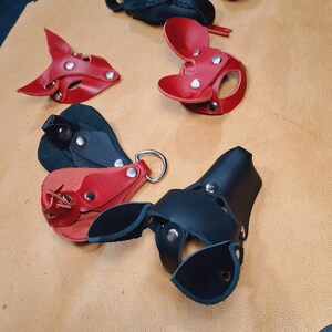 May include: A collection of handcrafted leather animal-shaped masks and keychains in red and black. The items feature silver-toned metal rivets and D-ring hardware, showcasing various ear shapes and snout designs constructed from layered leather pieces.