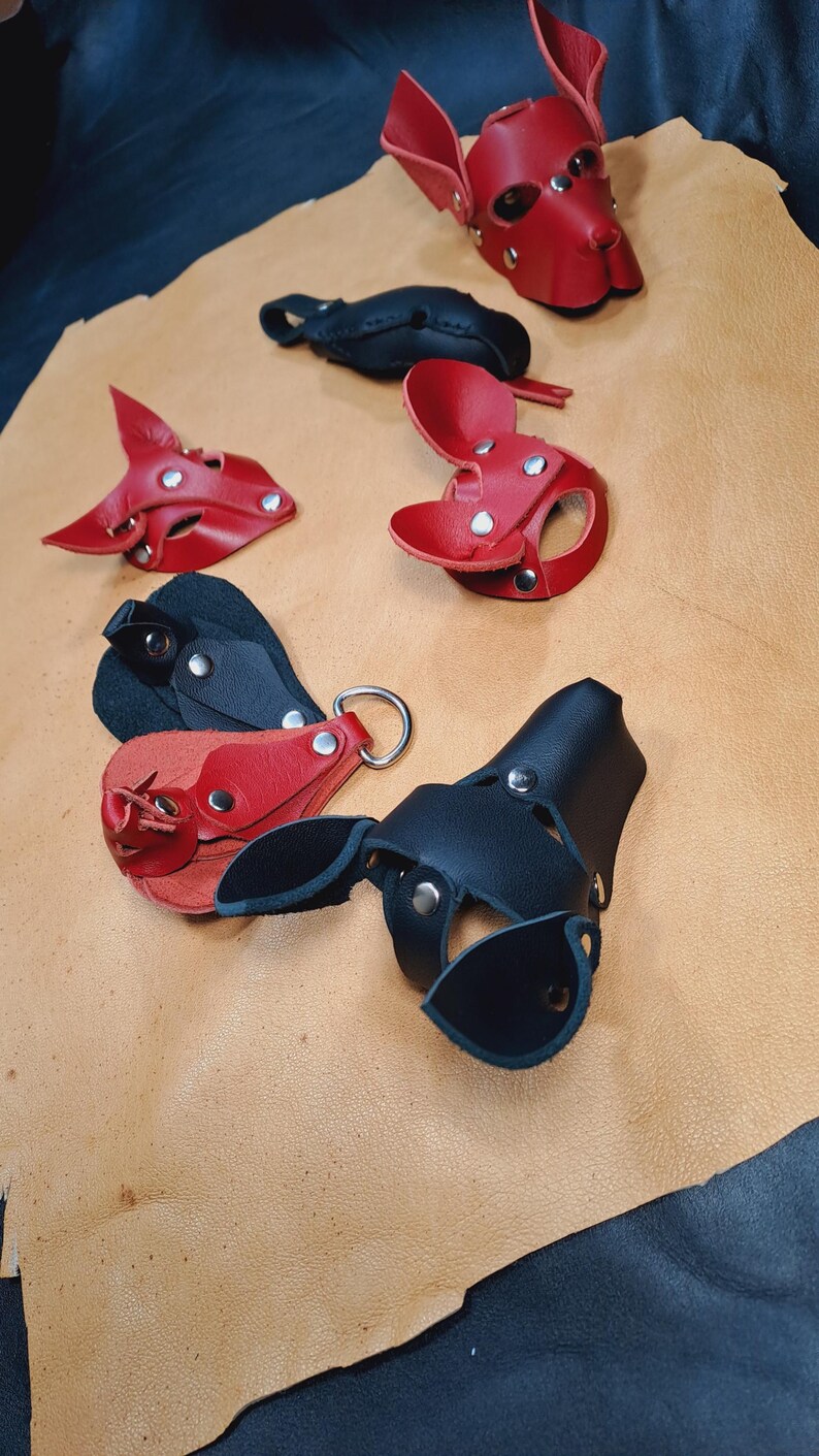 May include: A collection of handcrafted leather animal-shaped masks and keychains in red and black. The items feature silver-toned metal rivets and D-ring hardware, showcasing various ear shapes and snout designs constructed from layered leather pieces.