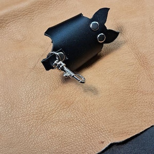 May include: A black leather accessory shaped like a bat with two silver-toned metal rivets and a silver-toned metal lobster clasp attached to a small loop. The item features pointed ears and a curved body design made of smooth black leather.