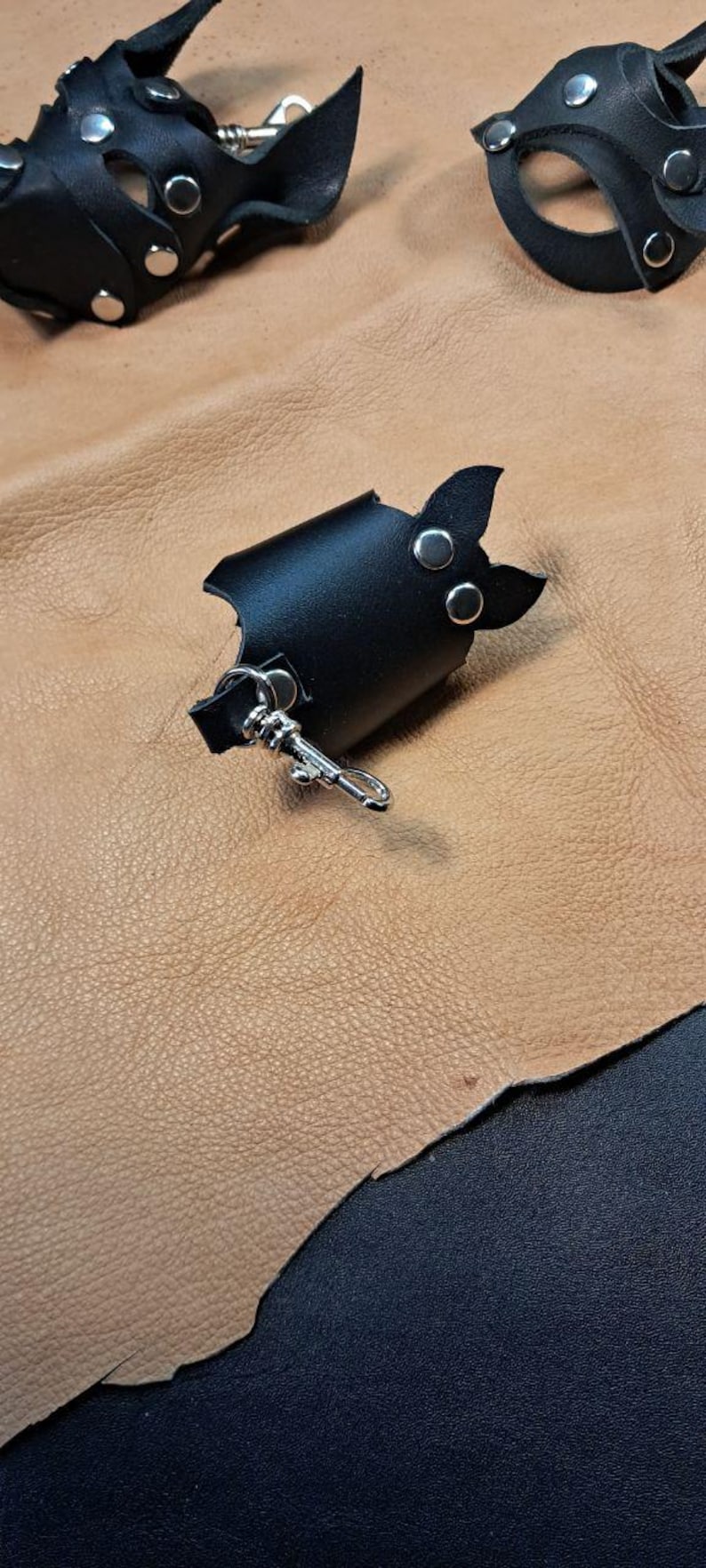 May include: A black leather accessory shaped like a bat with two silver-toned metal rivets and a silver-toned metal lobster clasp attached to a small loop. The item features pointed ears and a curved body design made of smooth black leather.