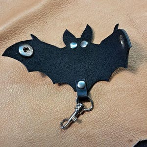 May include: A black leather bat-shaped keychain featuring silver-toned metal snaps for the eyes and wings, along with a silver-toned metal lobster clasp attached to the bottom. The design includes a central snap closure and detailed wing cutouts.