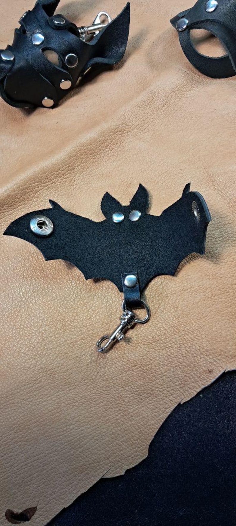 May include: A black leather bat-shaped keychain featuring silver-toned metal snaps for the eyes and wings, along with a silver-toned metal lobster clasp attached to the bottom. The design includes a central snap closure and detailed wing cutouts.
