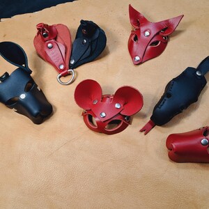 May include: A collection of six leather animal-themed masks and accessories in red and black. The items feature pointed ears, rounded ears, and snout-like shapes, all constructed with visible metal rivets and snaps for adjustable fastening.