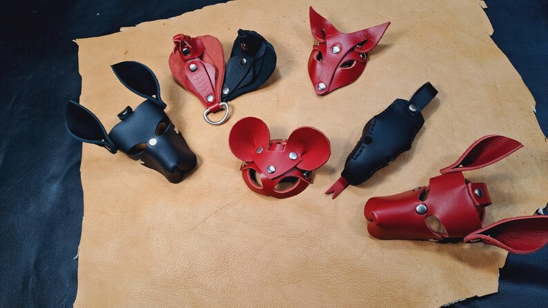 May include: A collection of six leather animal-themed masks and accessories in red and black. The items feature pointed ears, rounded ears, and snout-like shapes, all constructed with visible metal rivets and snaps for adjustable fastening.