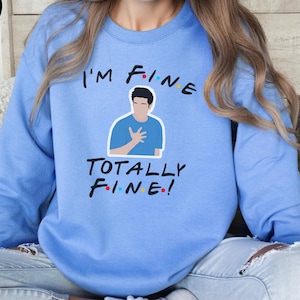Może przedstawiać: Błękitna bluza z napisem "I'M FINE" nad ilustracją osoby i "TOTALLY FINE!" poniżej. Bluza jest w jednolitym, jasnoniebieskim kolorze. Tekst i ilustracja są w kolorze czarnym, białym i czerwonym.