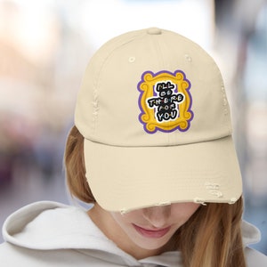 K&ouml;nnte beinhalten: Eine beige, abgenutzte Baseballkappe mit einem gestickten Aufn&auml;her in Gelb und Lila, der den Text "I'll be there for you" in einem wei&szlig;en Rahmen zeigt.