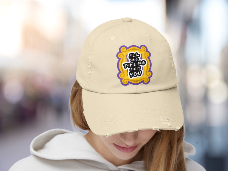 K&ouml;nnte beinhalten: Eine beige, abgenutzte Baseballkappe mit einem gestickten Aufn&auml;her in Gelb und Lila, der den Text "I'll be there for you" in einem wei&szlig;en Rahmen zeigt.