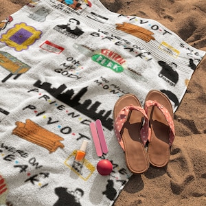 May include: White beach towel with a Friends TV show theme. The towel features various iconic images and quotes from the show, including the iconic "Pivot" scene, the Central Perk logo, and the phrase "We were on a break!" The towel is lying on a sandy beach.
