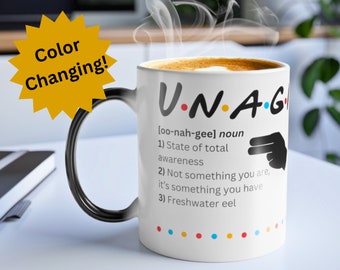 May include: A white ceramic mug with a black handle. The mug has a color-changing design that reveals a black and white illustration of a hand pointing to the text "UNAG" with the definition "[oo-nah-gee] noun 1) State of total awareness 2) Not something you are, it's something you have 3) Freshwater eel".