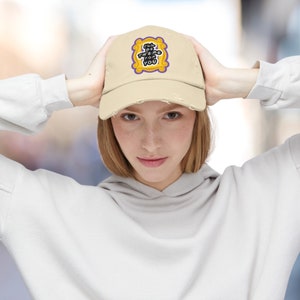 K&ouml;nnte beinhalten: Eine beige Baseballkappe mit abgenutztem Look. Die Vorderseite der Kappe zeigt einen gestickten Aufn&auml;her in Gelb und Lila mit dem Text "I'll be there for you" in einem lila Rahmen.